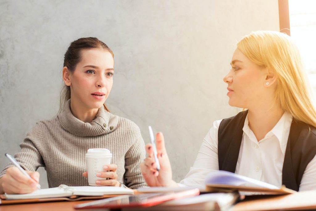 zwei Frauen sitzen mit Notizbüchern an einem Tisch