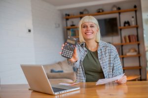 Studentin sitzt lächelnd am Laptop und hält Taschenrechner in die Kamera