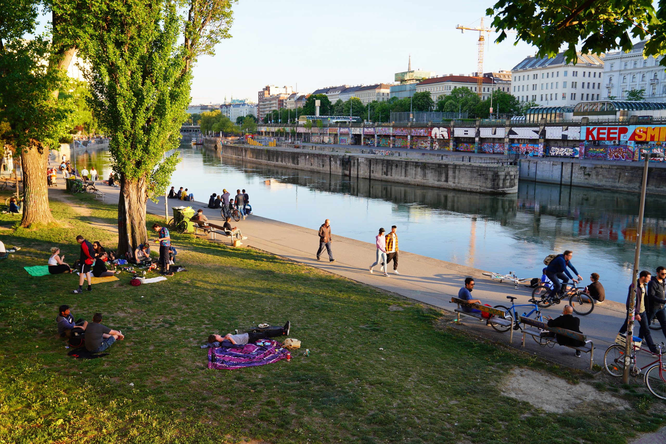An einem Frühlingstag spazieren die Leute den sonnigen Donaukanal entlang, manche liegen im Gras