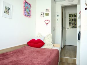 Ausschnitt eines Einzelzimmers in einer WG, Bett mit roter Decke und mehreren Zierpölstern, ein großer roter Kussmund Zierpolster in der Mitte des Bildes. Die Tür in den Vorraum ist offen, man sieht auf die Klo- und Badezimmertüre. an der Wand hängen Postkarten, Bilder und ein abstraktes buntes Bild welches ein Huhn abbildet. rechts ist zu erkennen, dass ein Regal an der Wand steht in dem eine schwarze Flasche steht