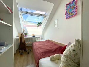 Einzelzimmer mit Einzelbett rechts und roter Tagesdecke. Ein schräges offenes Dachfenster. Am Fenstergriff hängt eine Pflanze. Darunter ein Schreibtisch mit diversen Sachen darauf. Vor dem Tisch steht ein Schreibtischsessel an dem eine Tasche hängt.